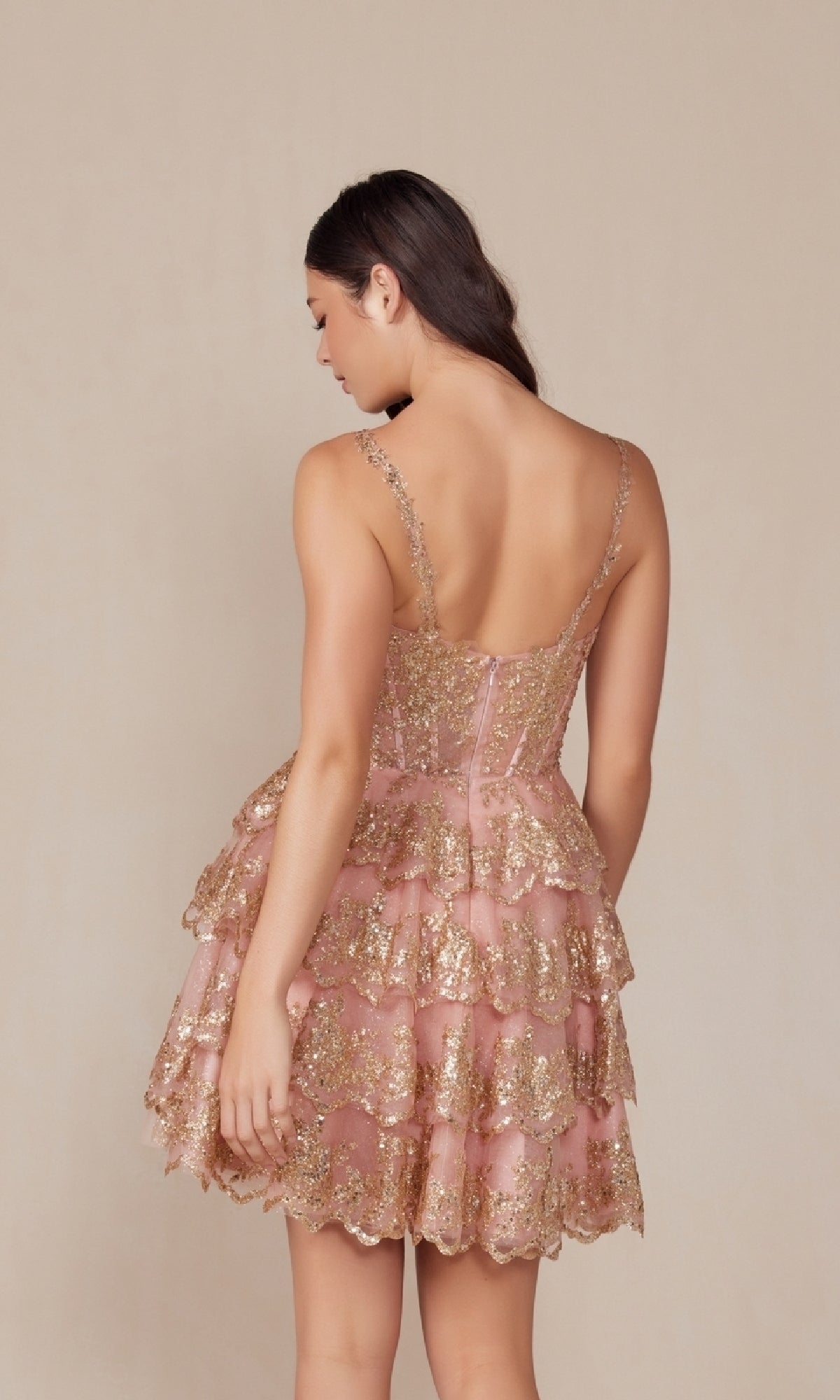 A woman with long brown hair faces away, wearing the sleeveless, tiered pink Nox Anabel E914 Sequin Glitter Homecoming Dress with gold glitter accents, set against a plain beige background.